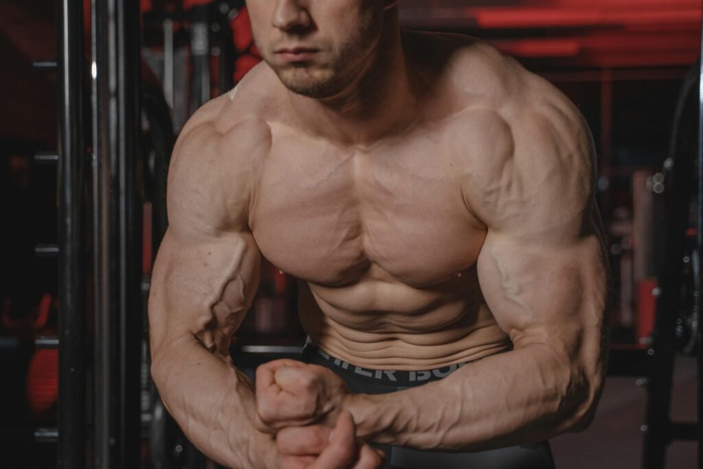 Shirtless man displaying muscular physique with focus on biceps and abs in a gym.